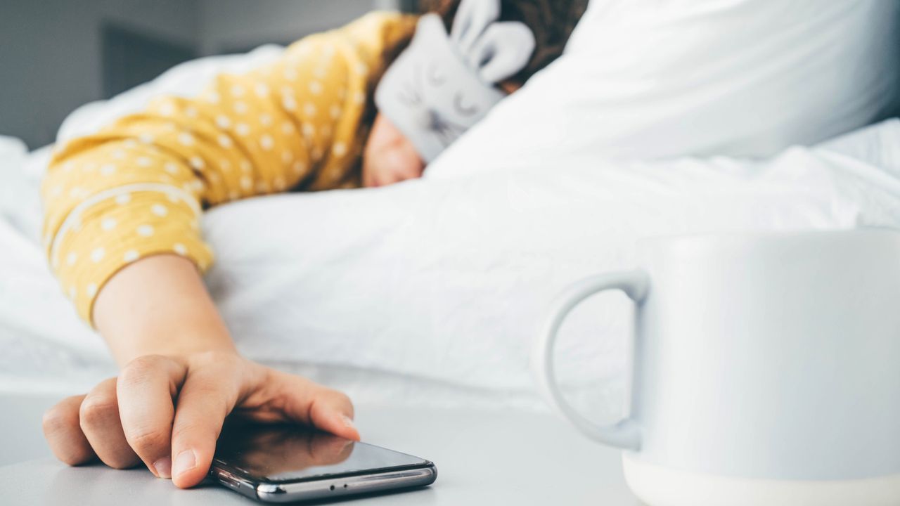 Woman wearing sleep mask in bed reaching out to turn off phone alarm