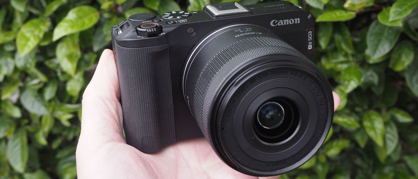 Canon EOS R50 V held in reviewer&#039;s hand against foliage backdrop.