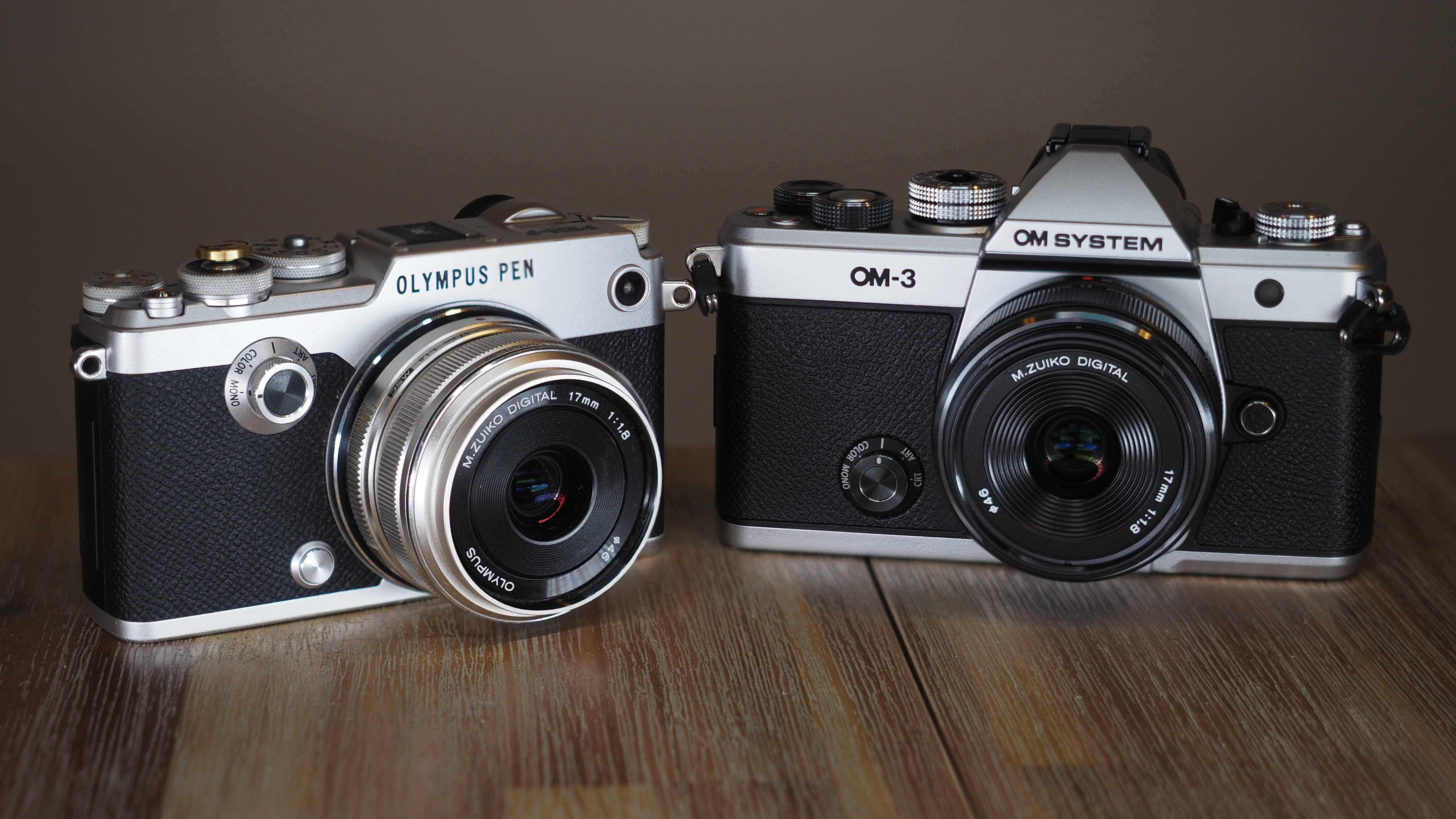 Ann Olympus PEN-F, next to an OM System OM-3, on a wooden surface