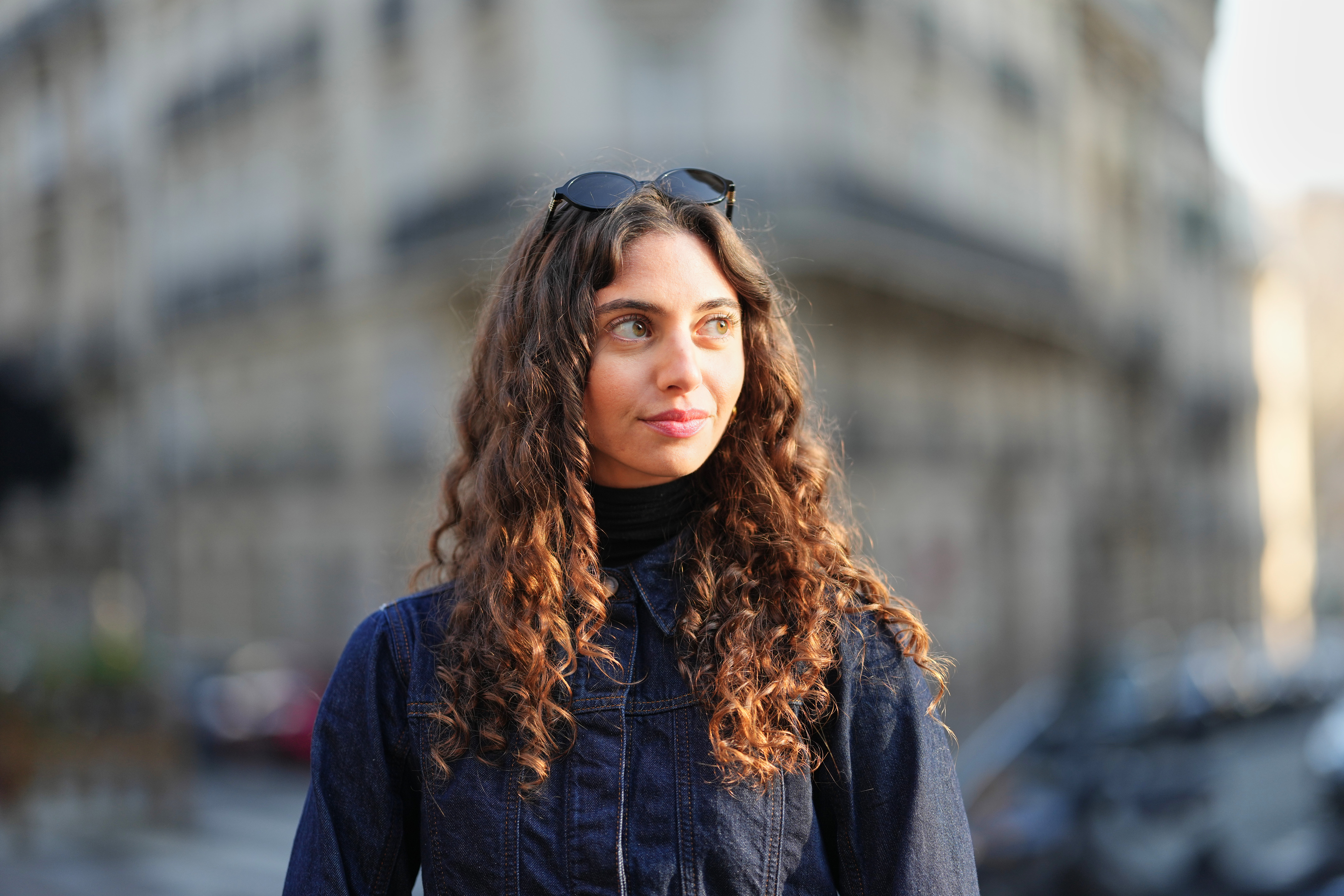 PARIS, FRANCE - DECEMBER 27: Amanda Derhy wears long curly / wavy brown hair worn loose with a center part, black oval sunglasses from Miu Miu, make-up is a natural complexion with defined brows and soft pink lipstick, a black ribbed turtleneck top layered under a dark indigo blue denim jacket with a pointed collar, during a street style fashion photo session, on December 27, 2025 in Paris, France. (Photo by Edward Berthelot/Getty Images)
