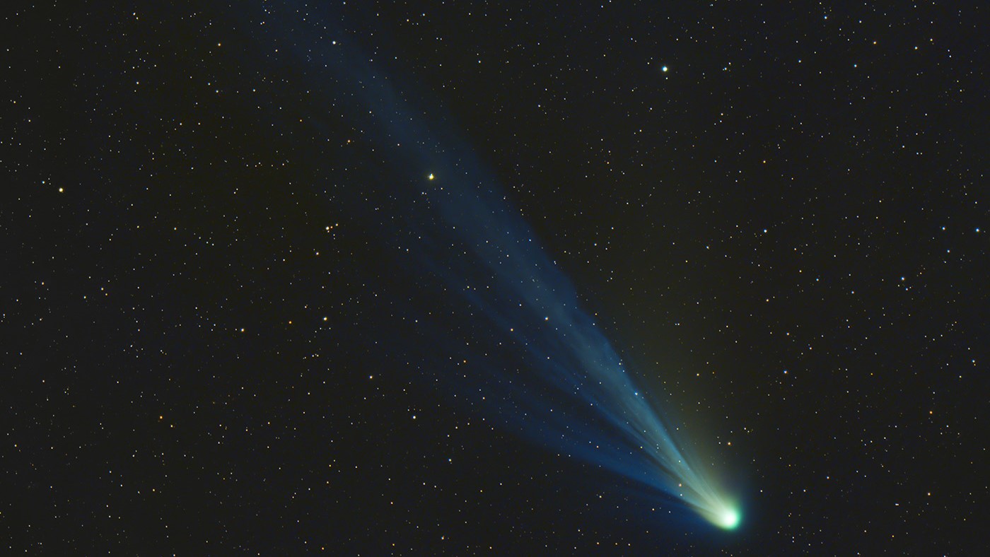 Comet 12P/Pons-Brooks appears as a bright green nucleus and a long blue tail streaking behind it against a background of stars.