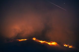 orange flames glowing from a black pit of clouds reflect off smoke in the sky as a shooting star streaks by the upper right.