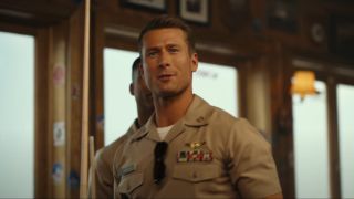 Glen Powell walking and talking with a pool cue in hand in Top Gun: Maverick.
