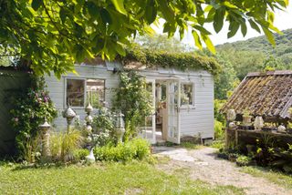 a rustic white outbuilding with lots of decor outside and plenty of plants and foliage in the garden