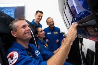four astronauts in blue flight suits sit near computer monitors