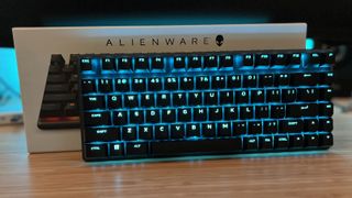 Alienware Pro Wireless gaming keyboard with blue RGB on, leaning against packaging on a wooden desk