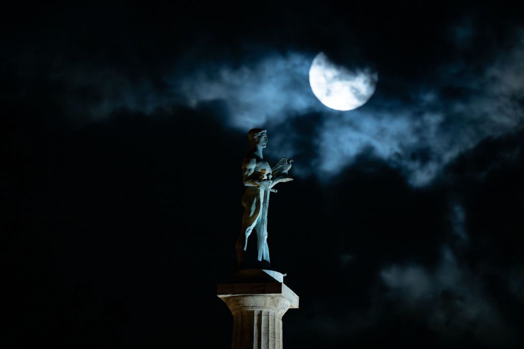 La luna azul gigante está parcialmente obstruida por nubes en esta imagen de mal humor que la muestra elevándose sobre un monumento a una persona parada en un poste alto.