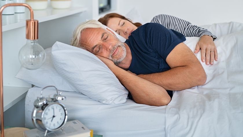 This image shows a senior man and woman sleeping in bed. The man lies closest to the camera, while the women is in the background. They're both facing the camera. In the foreground is a lamp and an alarm clock