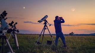 Man uses binoculars to look at the night sky as the sun sets and telescopes are on tripods around him