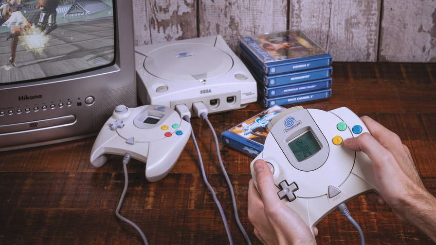 A photo showing someone holding the controller for a Sega Dreamcast, with the console sat on a table next to a small collection of games, a second controller, and a TV showing a game running.