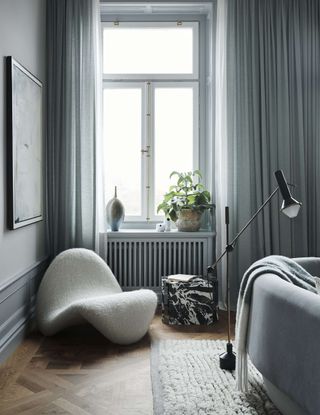 armchair and marble side table styled in a living room next to a window with curtains, a radiator, with a pot plant and ceramic vase on the windowsill. in the foreground, a floor lamp leans over a curved sofa