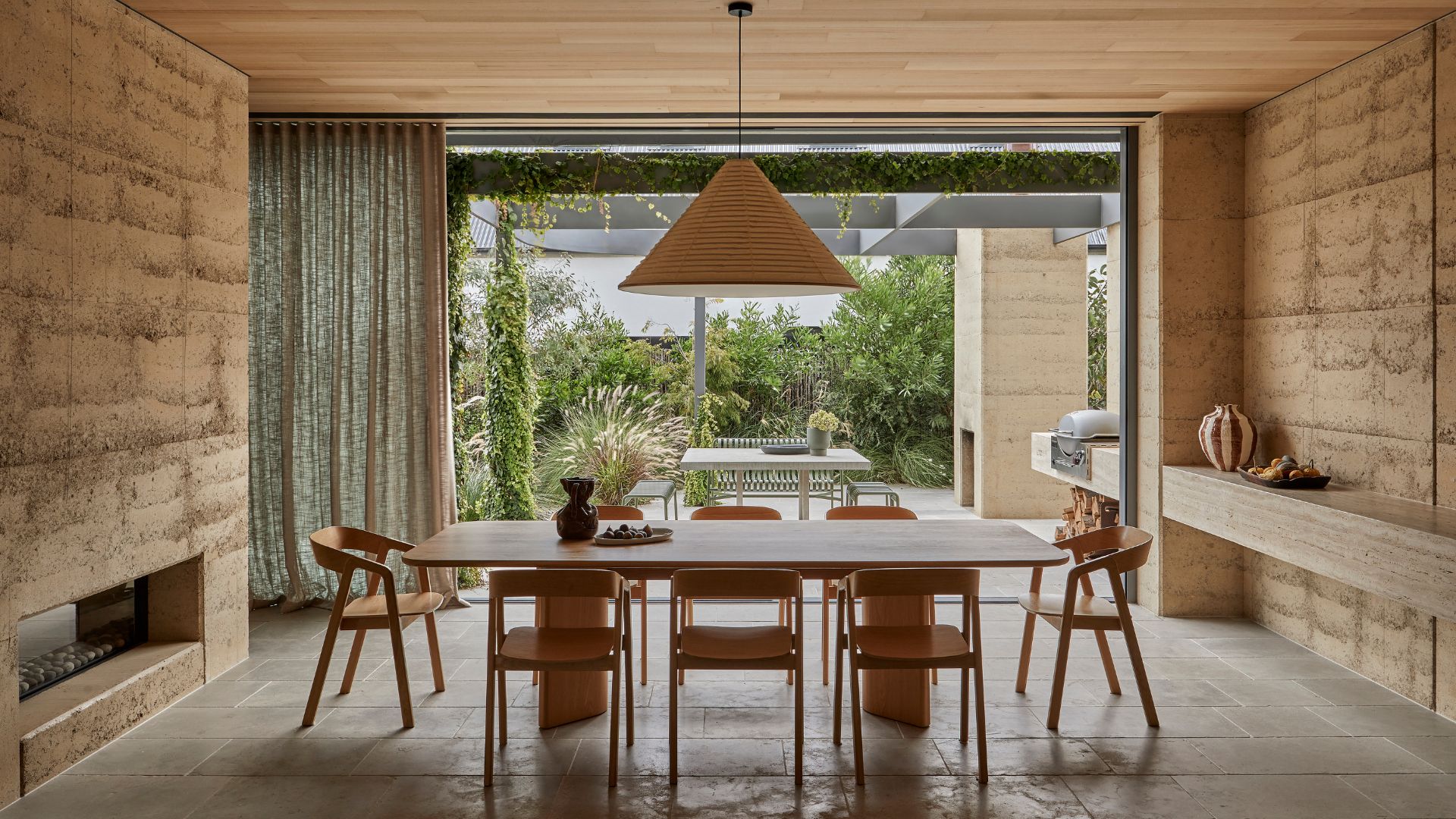 A dining room with a wood dining set, a conical lantern, and a large sliding glass door looking out onto an outdoor dining room and grill