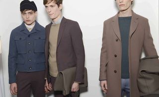 Three male models wearing looks from Gucci's collection. They are wearing blue, brown and dark beige trousers, jackets, sweaters and a coat. One model is wearing a dark hat and the other two models are holding bags