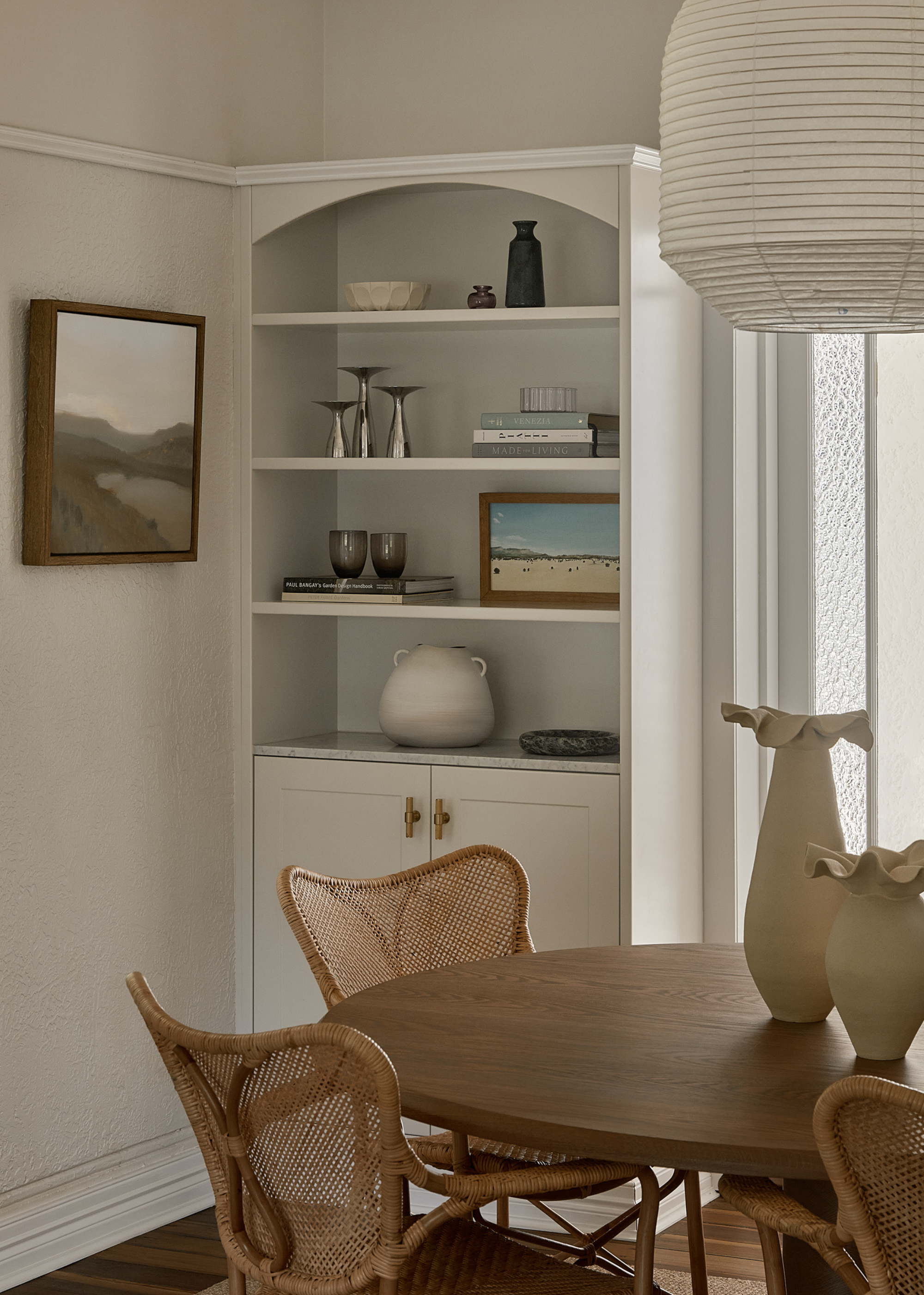 A white dining room with a shelf of books, knick knacks, glassware, and vases by a wood dining table, rattan chairs, and a rice paper pendant lamp