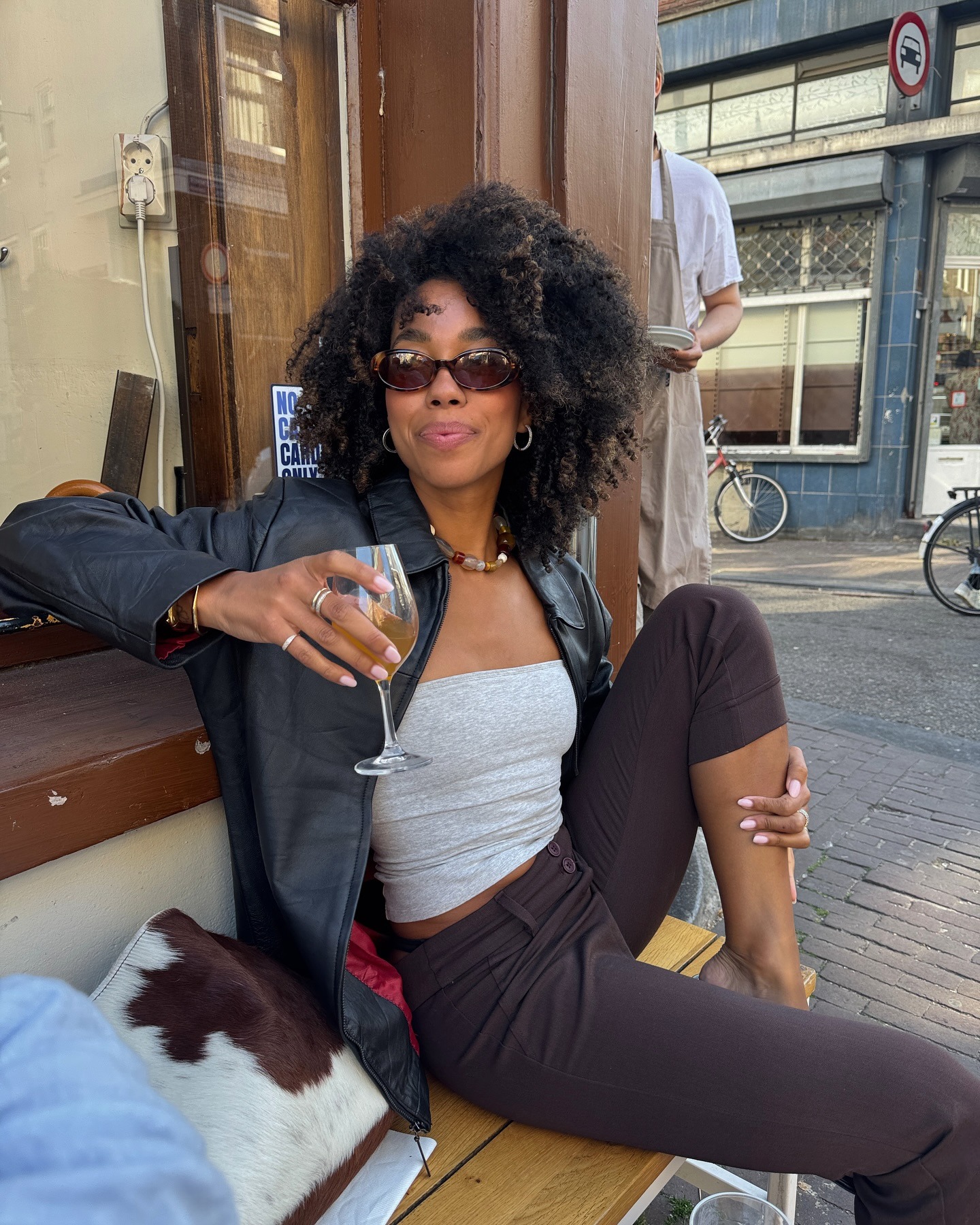 amaka.hamelijnck wears a pedal pusher outfit summer 2026. she wears brown capri's a grey tube top a black leather jacket and flip flops. She finishes the look with brown oval sunnies.