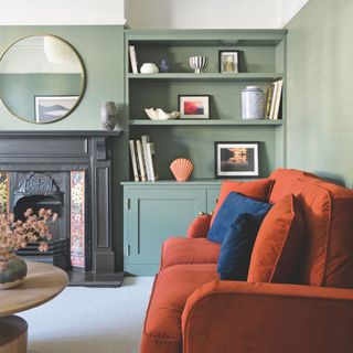 Pale green living room with a bright orange sofa and blue cushions. There's an old fireplace in the centre of the wall