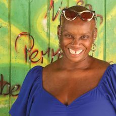 Andi Oliver smiling wearing a blue dress in front of a green wall
