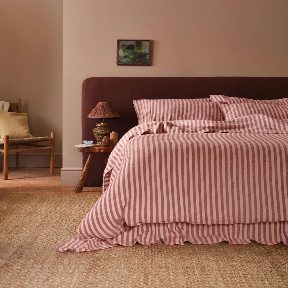 Image of a bed with a burgundy headboard and a pink and burgundy striped bedding set. The walls are color-drenched in a dusty pink color and there is a jute rug on the floor.