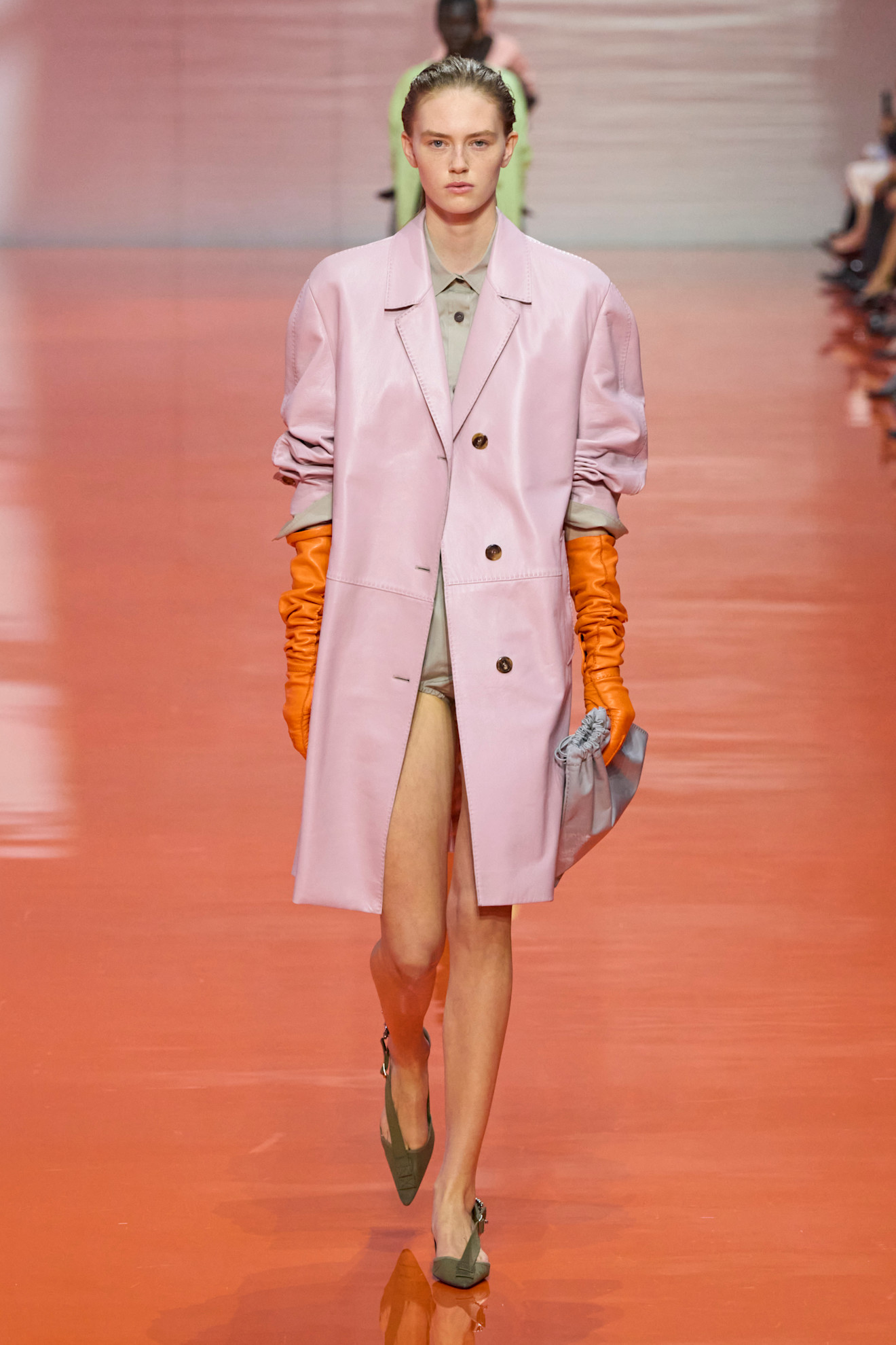 A model at the Prada SS26 show wearing a pink leather car coat with orange gloves.
