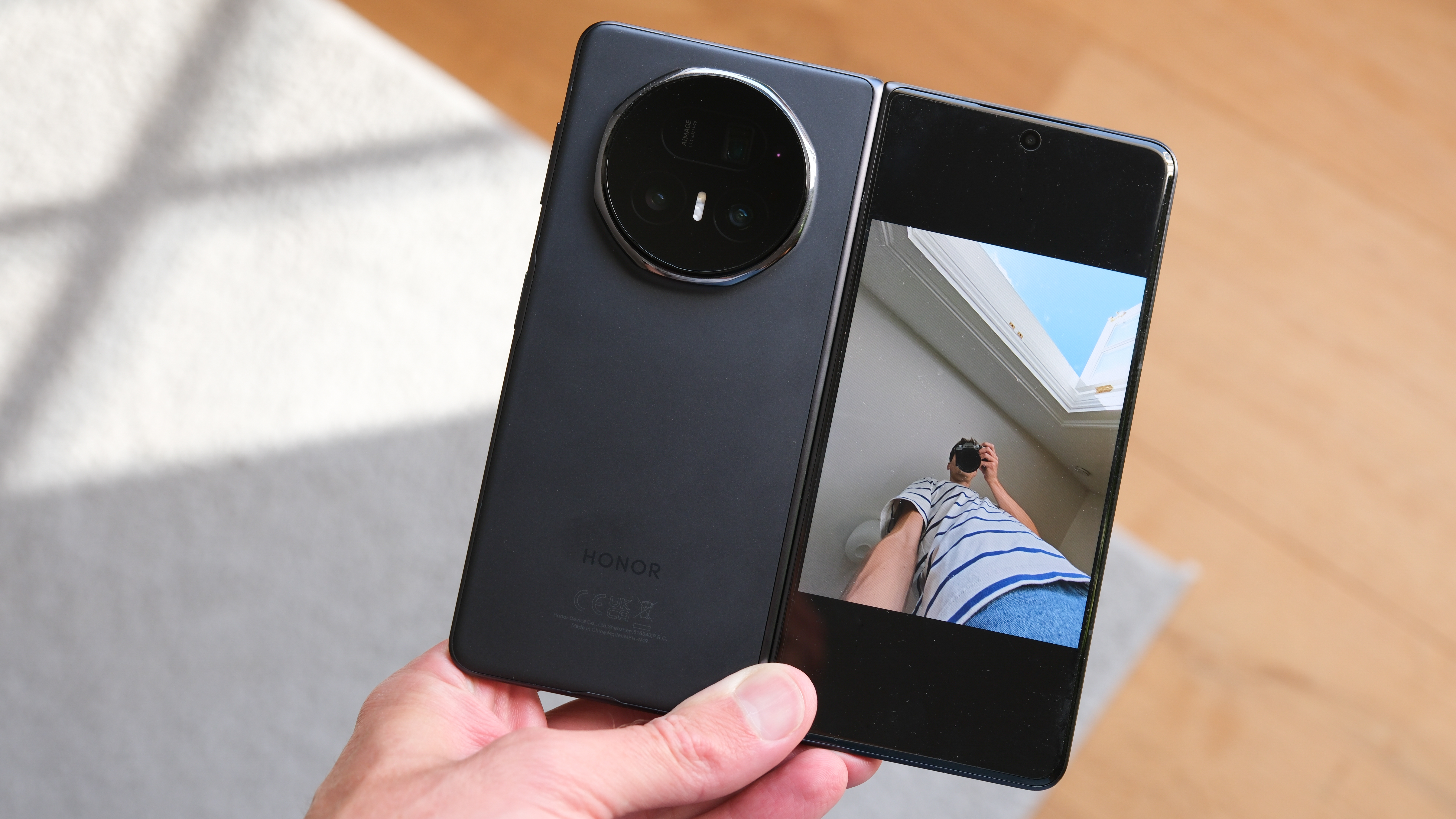 Honor Magic V5 folding phone held in a hand with the cover screen showing a person taking a picture of the phone