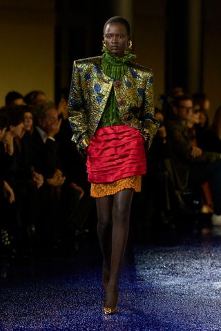 A model at Saint Laurent Spring 2025's runway show in a green top, green jacket, and red skirt.