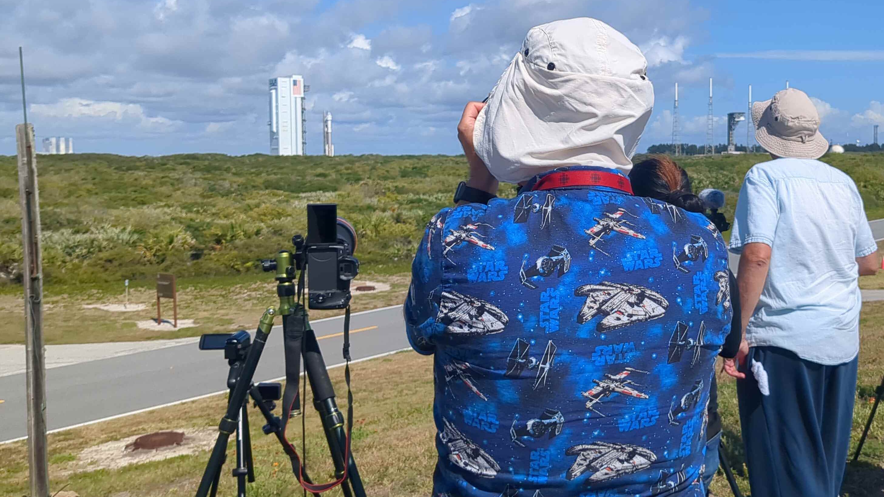 a person in a shirt with stars wars spacecraft facing a rocket and building, far in the background