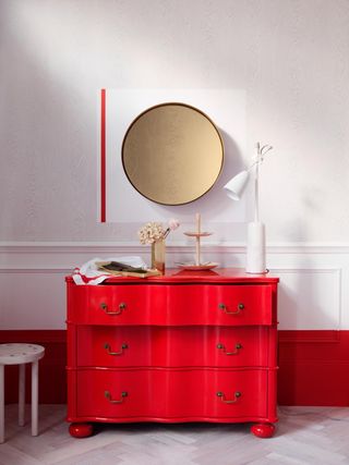 A white and red color-blocked wall with a bright red chest of drawers in front of it. There is a golden decorative circle hung above the chest and a white lamp sitting on it.