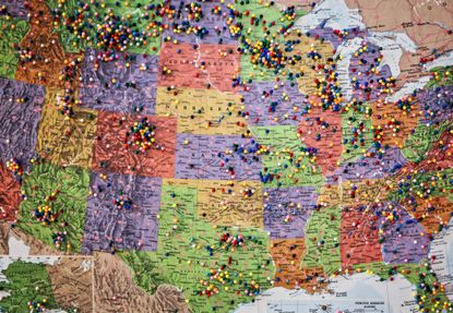 A paper map of the United States map hanging on a wall dotted with colorful pins marking destinations within 50 states