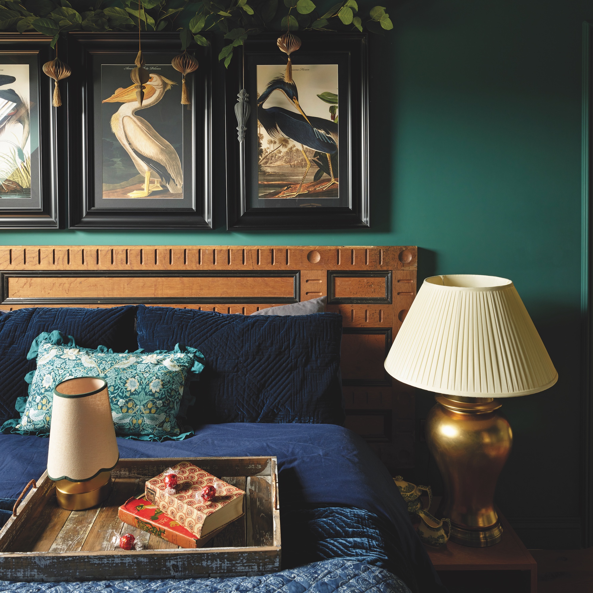 bedroom with dark green wall and double bed with navy bed linen, table lamp with brass base and cream shade, and prints of birds on wall