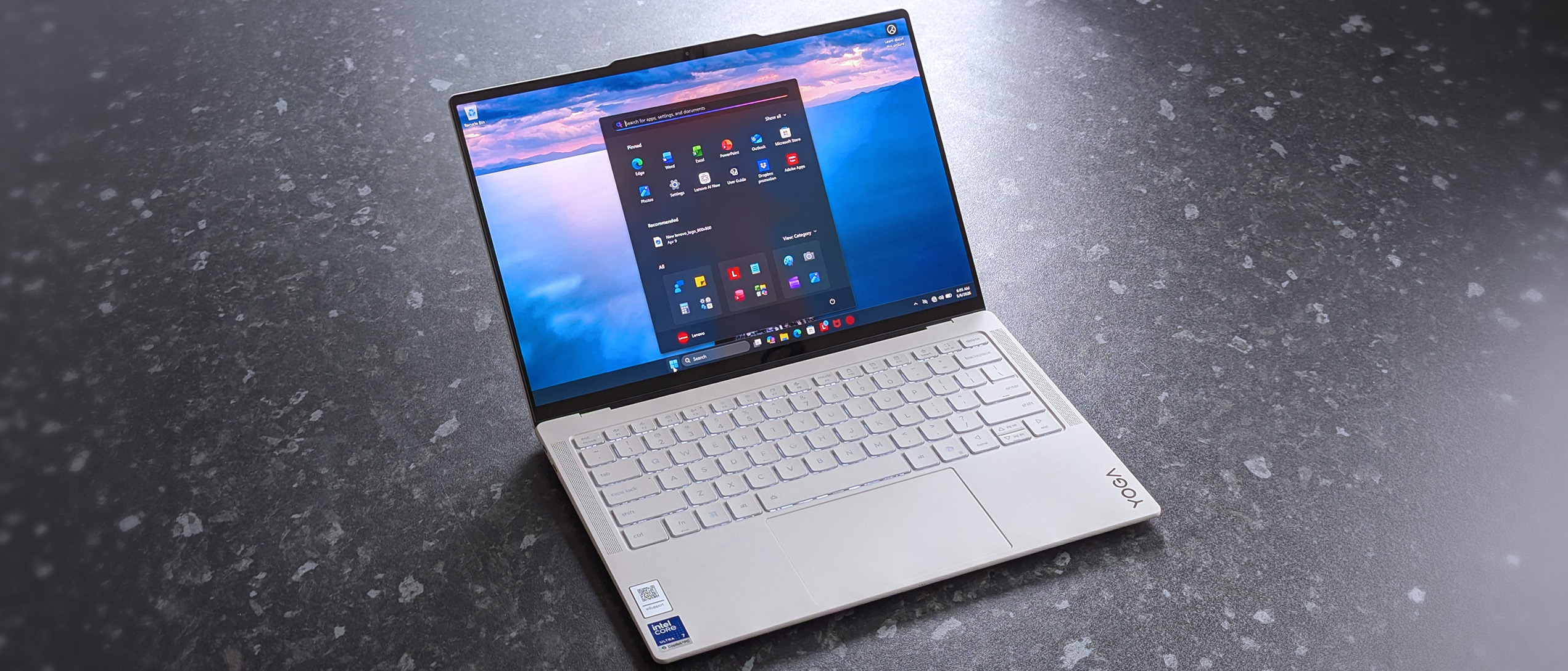 A sleek Lenovo Yoga Slim 7i Ultra (Gen 11) Aura Edition laptop on a dark, textured surface. The screen displays a vibrant ocean background with an open app menu.