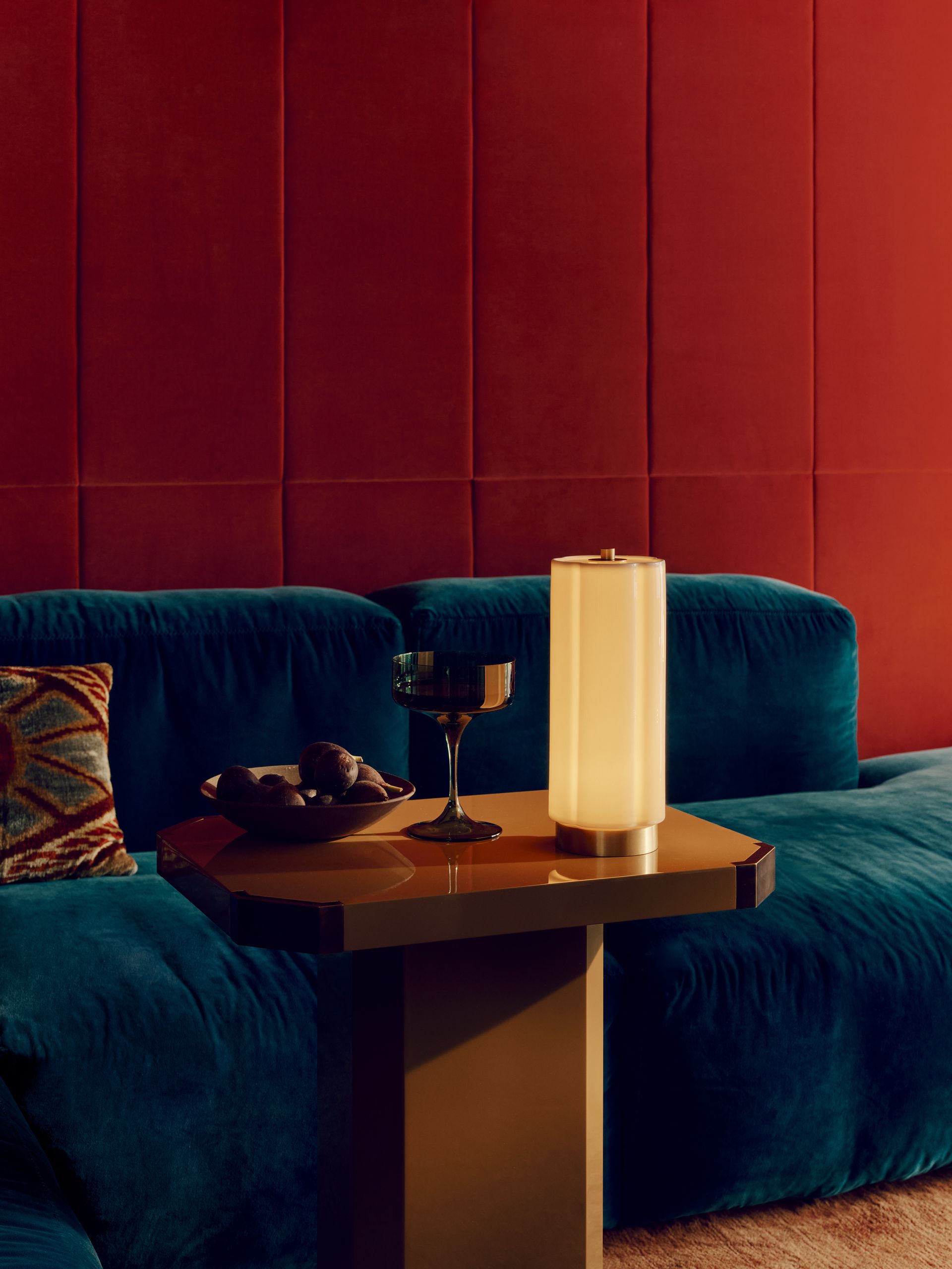 Image of a portable lamp on a lacquer side table in front of a blue velvet sofa in a red room.