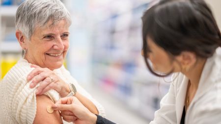 An older woman gets a circular band aid put on her by a woman with dark hair wearing a white lab coat.