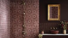 A deep purple/burgundy bathroom with an exposed brass shower against a paler purple/pink tiling and a bowl sink on a vanity below a gold-framed mirror and next to a vase of flowers