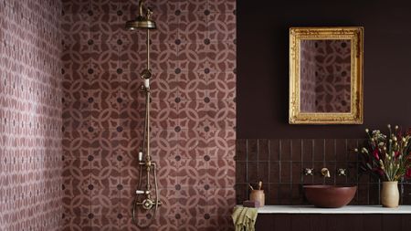 A deep purple/burgundy bathroom with an exposed brass shower against a paler purple/pink tiling and a bowl sink on a vanity below a gold-framed mirror and next to a vase of flowers