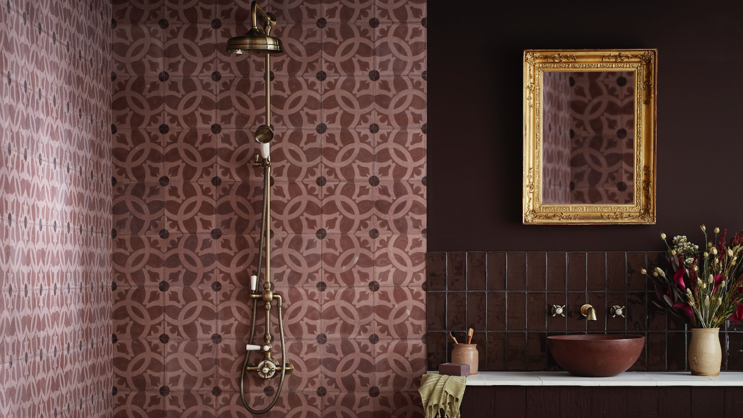 A deep purple/burgundy bathroom with an exposed brass shower against a paler purple/pink tiling and a bowl sink on a vanity below a gold-framed mirror and next to a vase of flowers