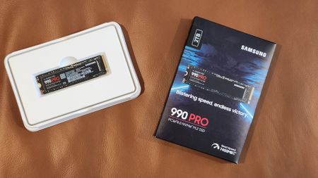 Samsung 990 Pro SSD on a brown surface, partially in a white case. The packaging highlights "2TB" and "Blistering speed, endless victory."