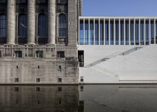 A side-on view of two buildings that are connected to each other. On the left, the Connection with Pergamon museum which has large pillars in grey. On the right, a white building with slender pillars and a stair case leading up to the upper level.