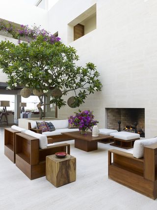 Modern indoor-outdoor entertaining area with matching bright walls and floors, fireplace and tree with wooden, cushioned sofa
