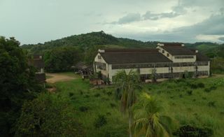 A still from a film by Juriaan Booij showing a large brown and off-white coloured building with multiple windows in Fordlandia during the day. The building is surrounded by hills and greenery