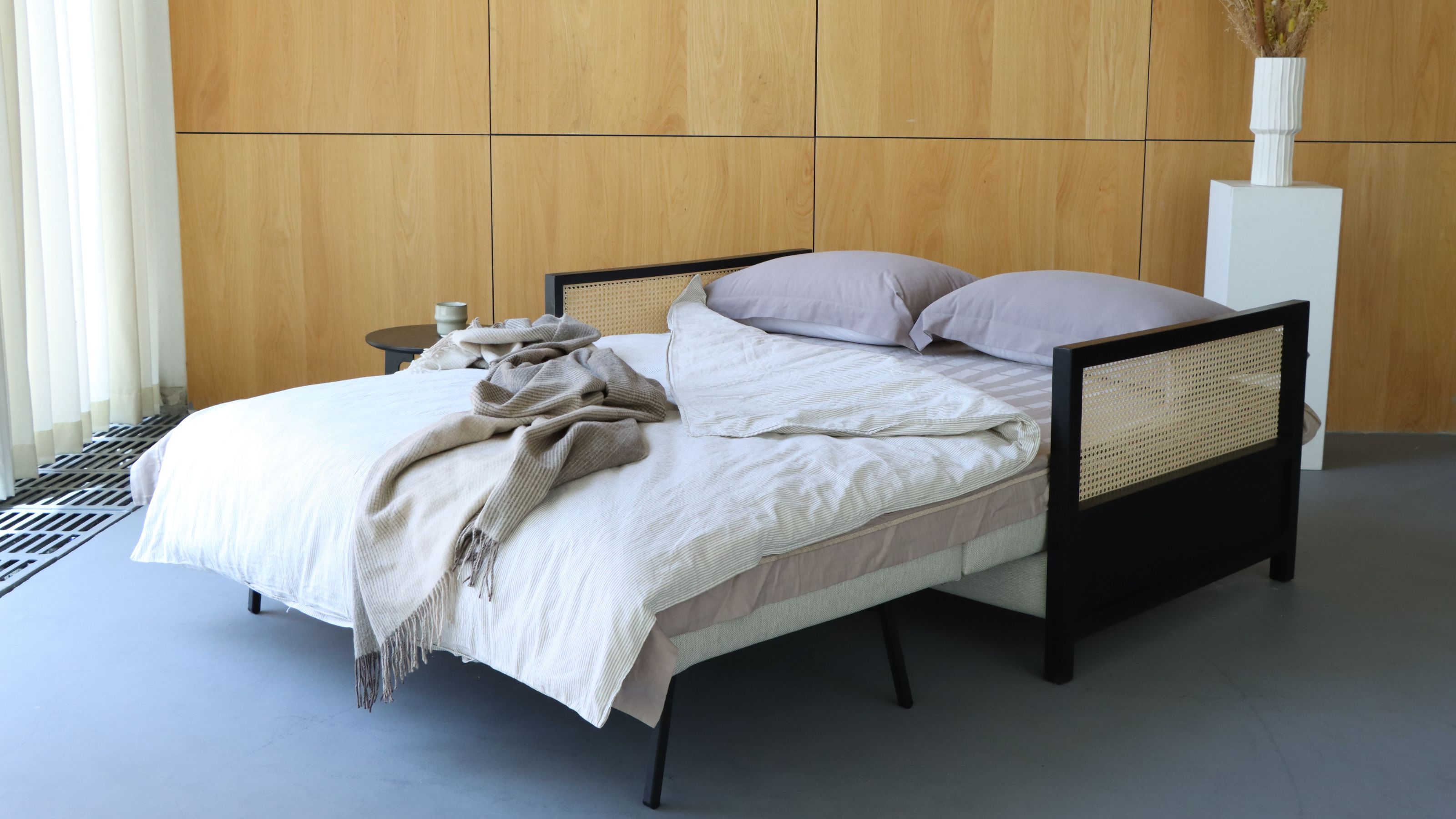 A sofa bed opened up with pillows and a blanket as well as a throw on top. There is also a view of a plant stand with a vase and flower inside of it