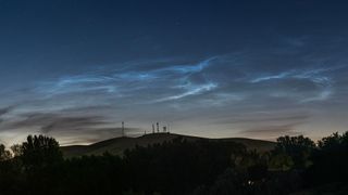 Silver-blue wispy clouds glow in a darkening evening sky above a hillside lined with trees.