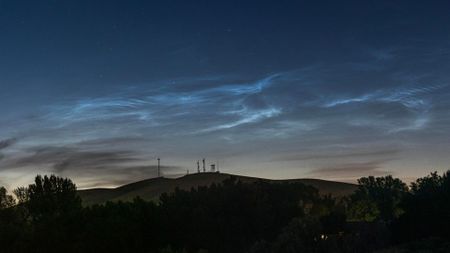 Silver-blue wispy clouds glow in a darkening evening sky above a hillside lined with trees.