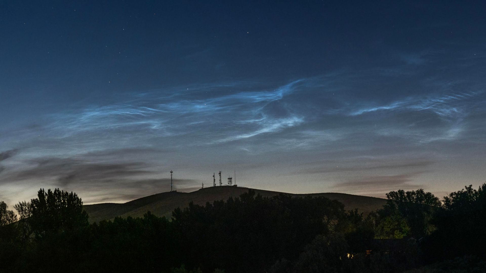 Silver-blue wispy clouds glow in a darkening evening sky above a hillside lined with trees.