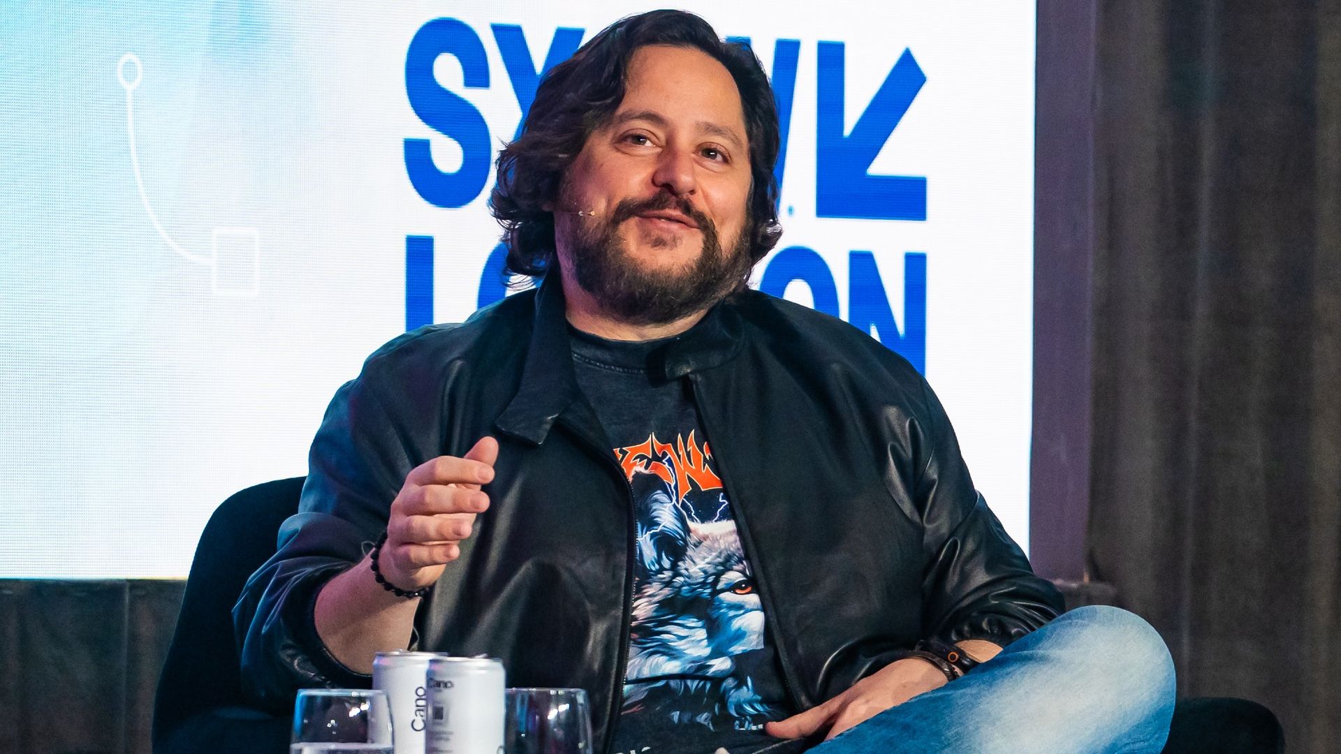 a man in a leather jacket and wolf t-shirt sits on a podium on a stage in front of a sign that reads 'sxsw london'