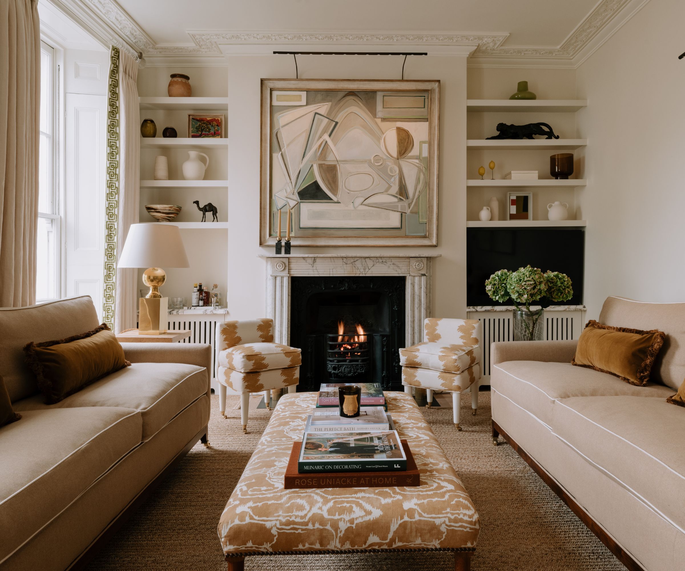 cozy modern meets traditional living room with a fireplace, alcove shelves, a marble fireplace surround, beige sofas with piping detail and a beige patterned ottoman