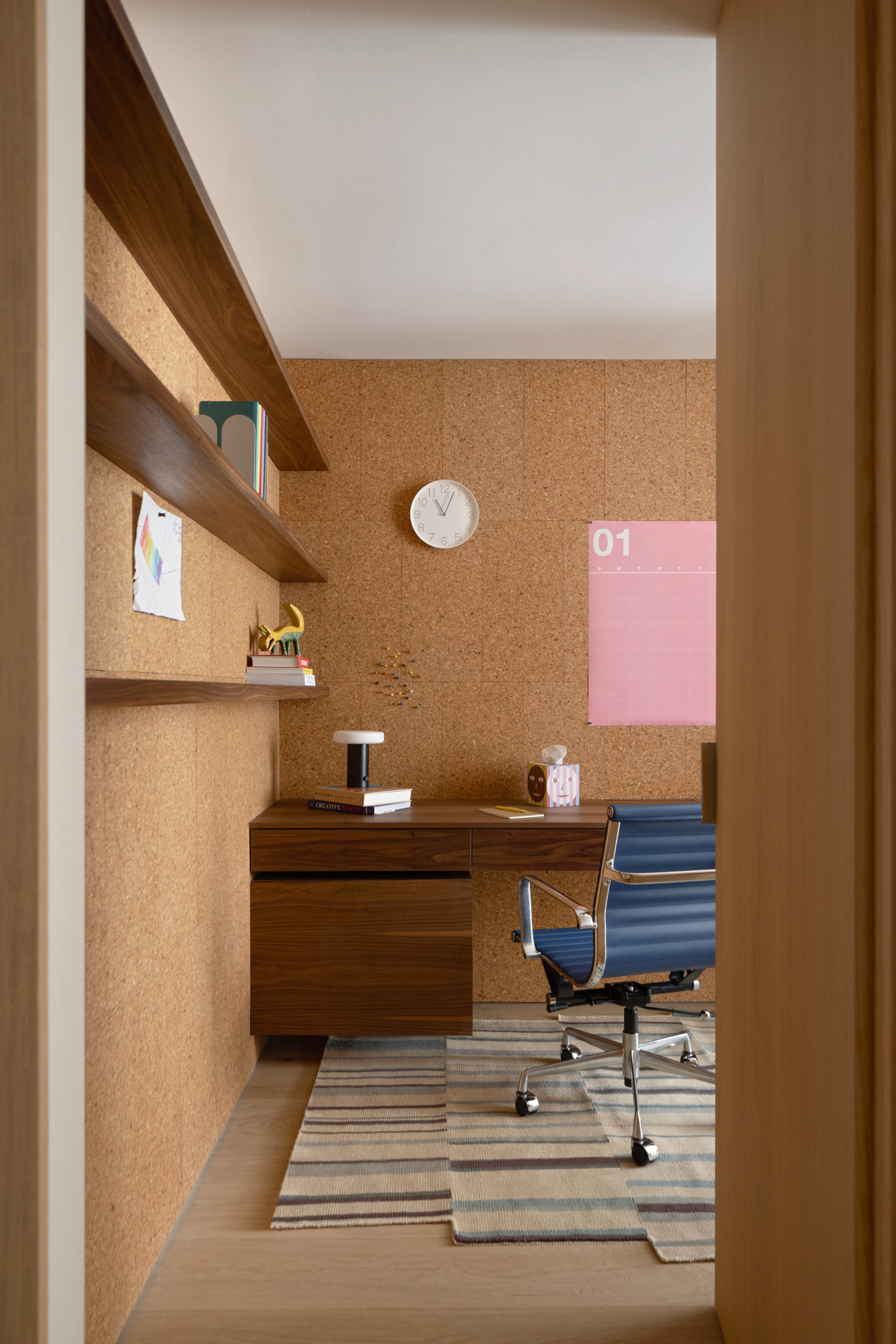 Home office with cork walls, wood shelving and desk, pink wall calendar and blue desk chair