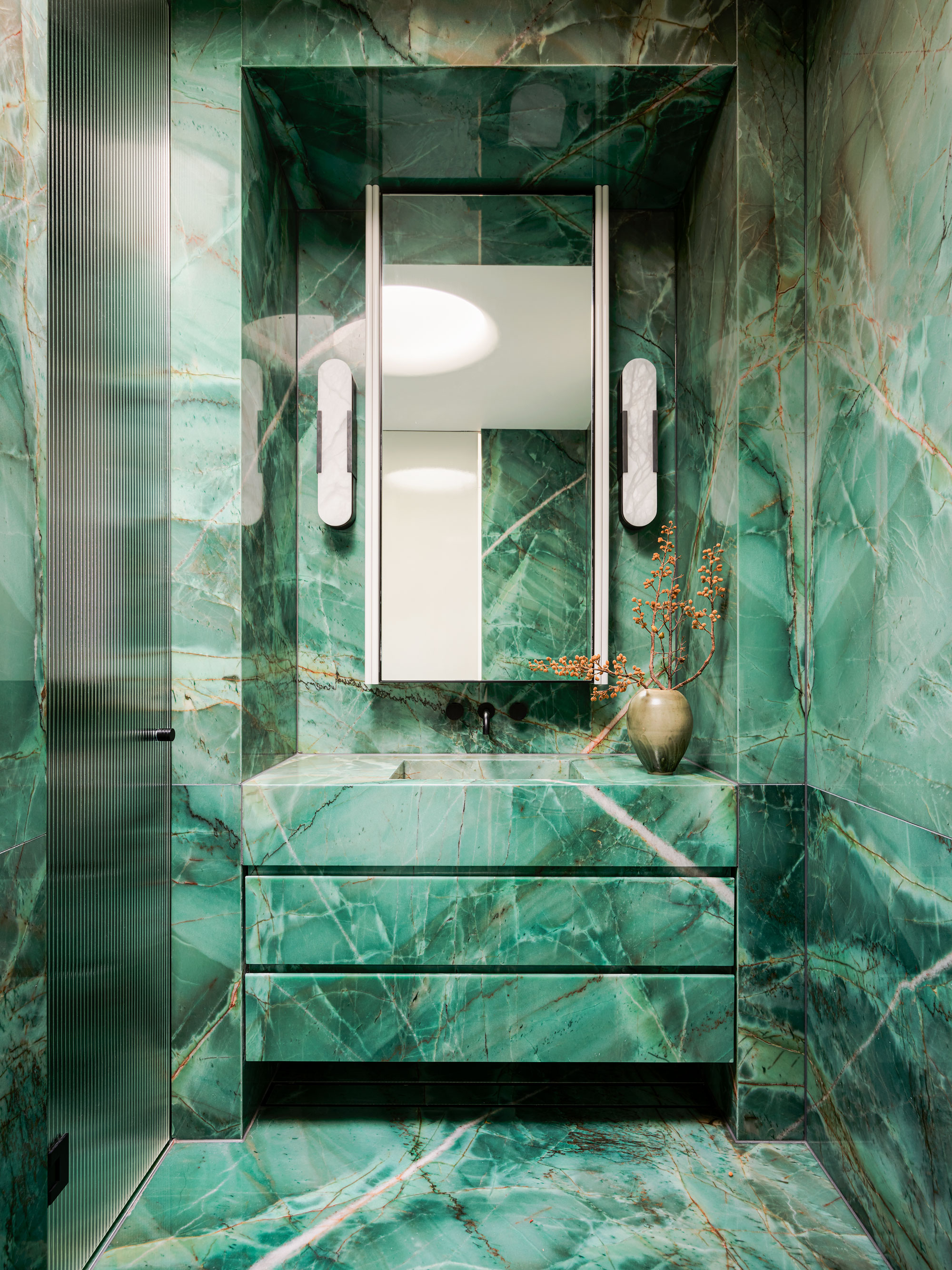 A jade stone drenched bathroom, with a view of the sink area and mirror, with the glass door to the shower to the right.