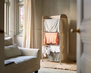 An image of the Eclipse™ 3-tier Ecru Clothes Drying Rack With Screen in a living room with clothes