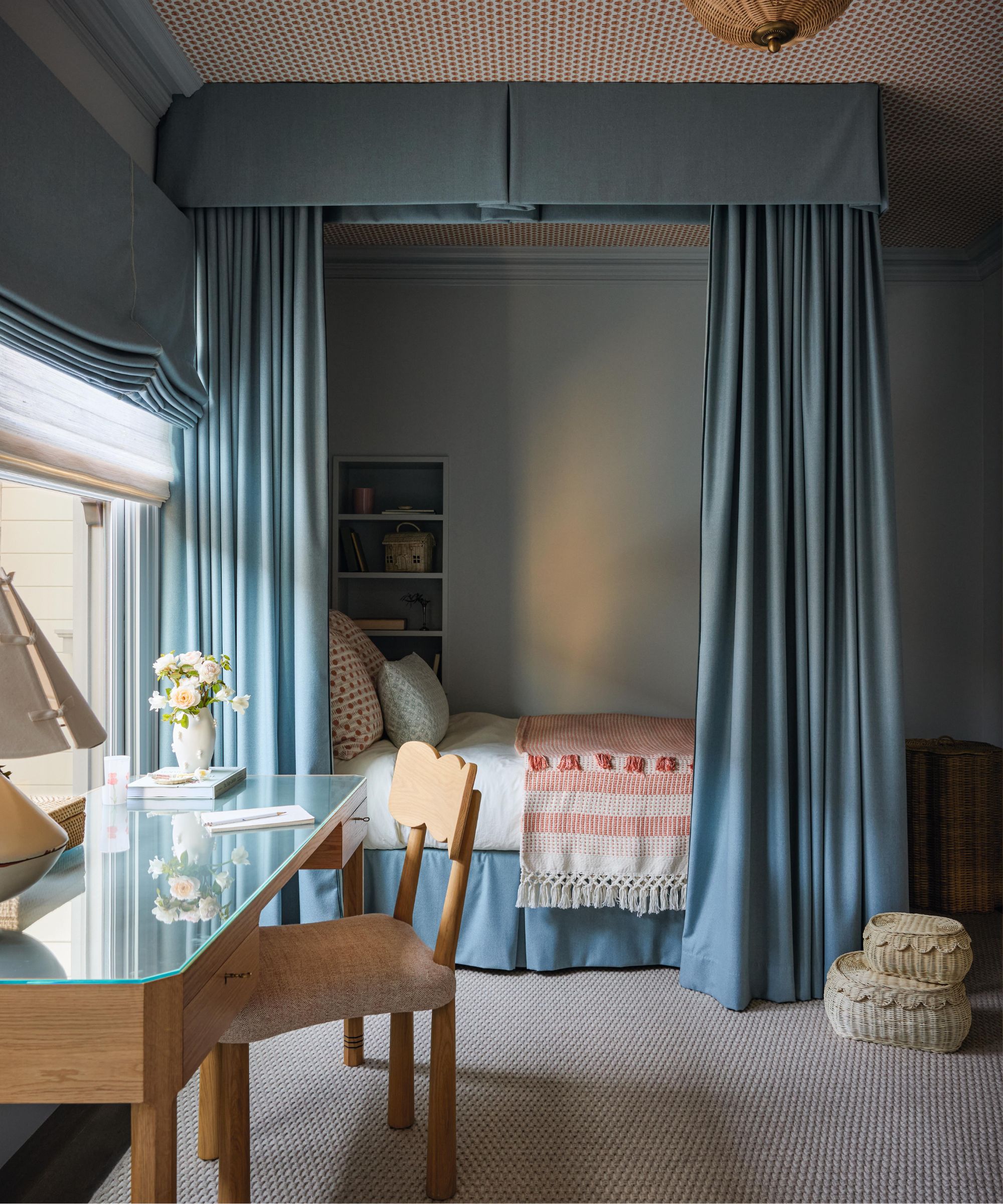 a girls bedroom with a patterned wallpapered ceiling, a blue draped bed nook and a small wooden desk under a window