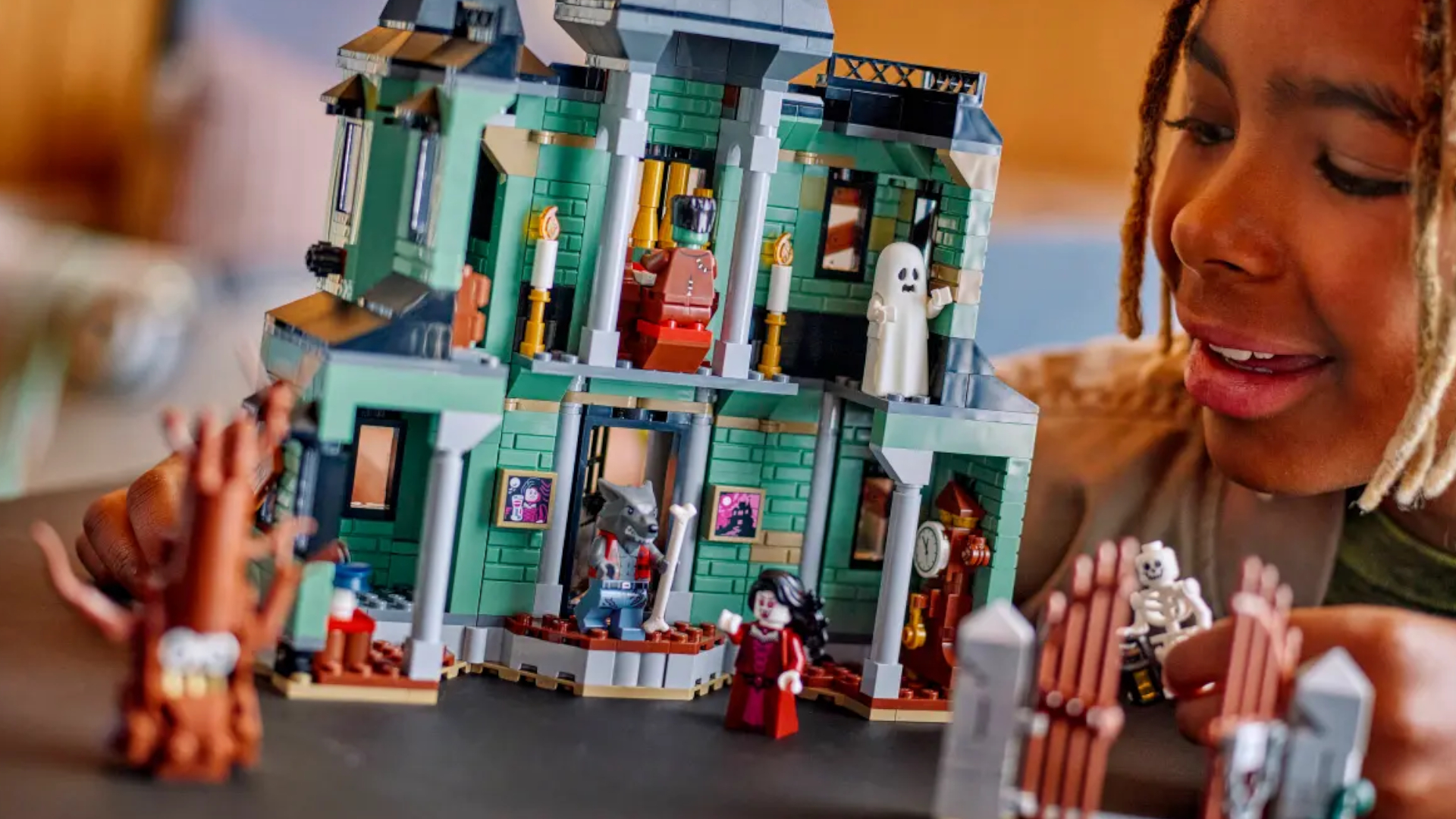 A child smiles at a green, spooky Lego house while placing a skeleton minifigure on a black surface
